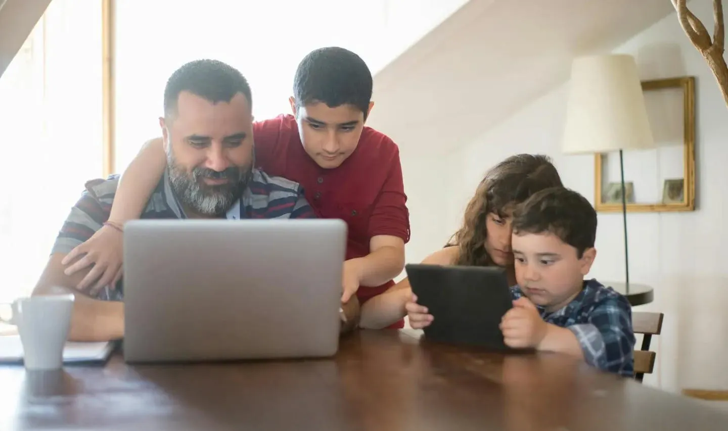 A father sitting at a table working on a laptop with his three young children with him