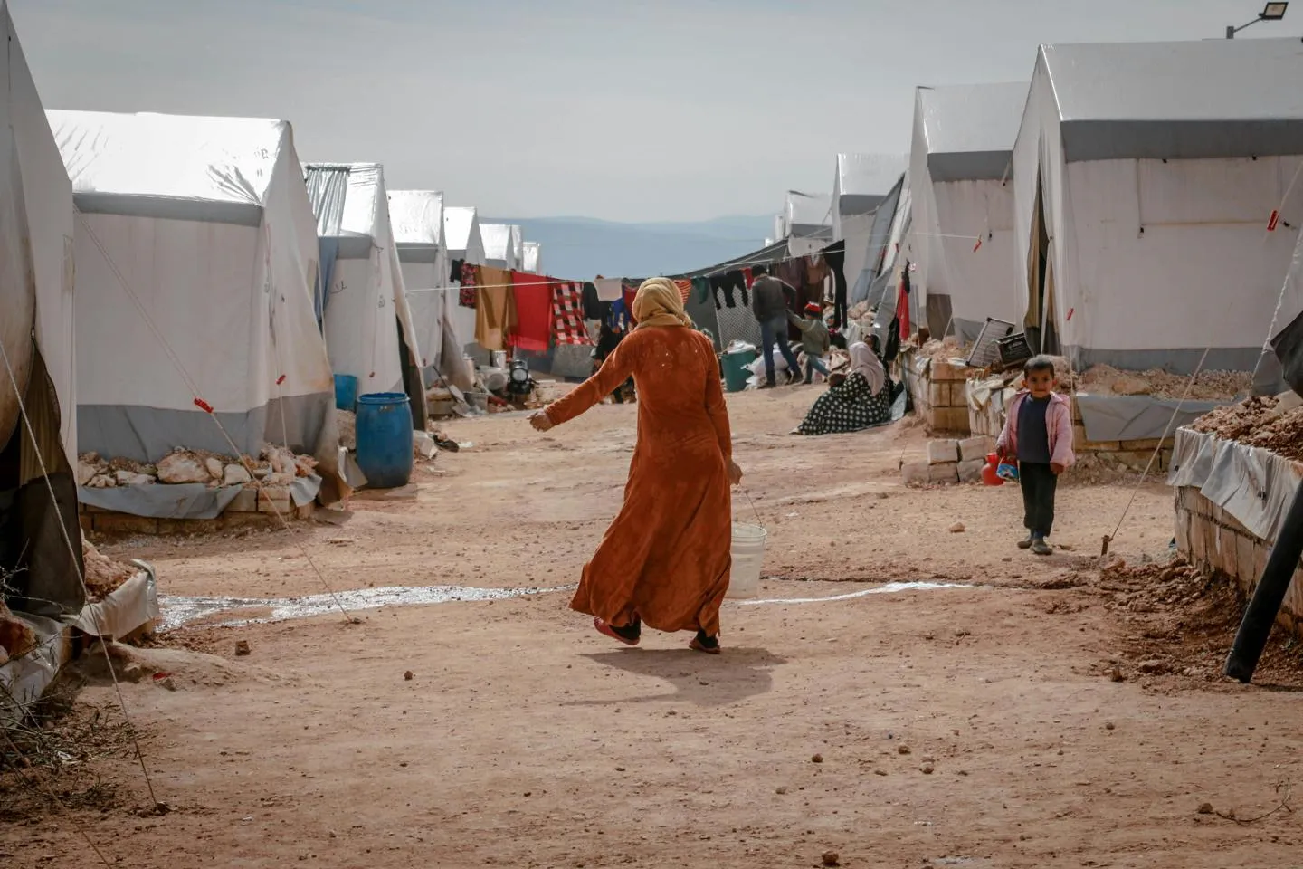 A woman walking through a refugee camp on a dirt track between two rows of white tents 